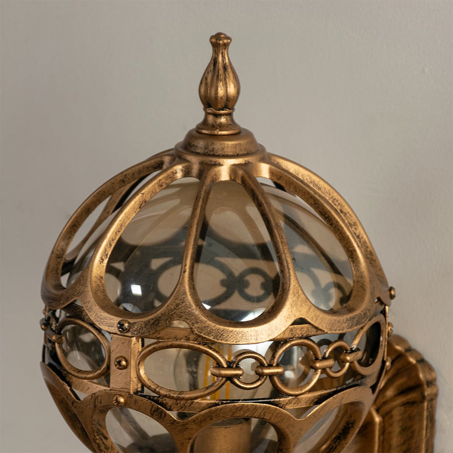 Closeup of gold metal outdoor wall lantern with dome glass design and ornate frame detailing for stylish decorative exterior lighting.