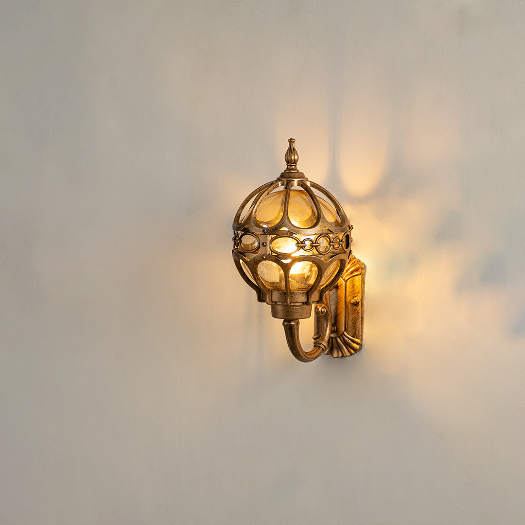 Antique brass outdoor wall sconce on white background featuring an ornate metal globe cage casting a warm radial shadow.