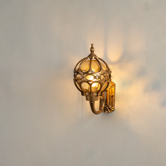 Antique brass outdoor wall sconce on white background featuring an ornate metal globe cage casting a warm radial shadow.