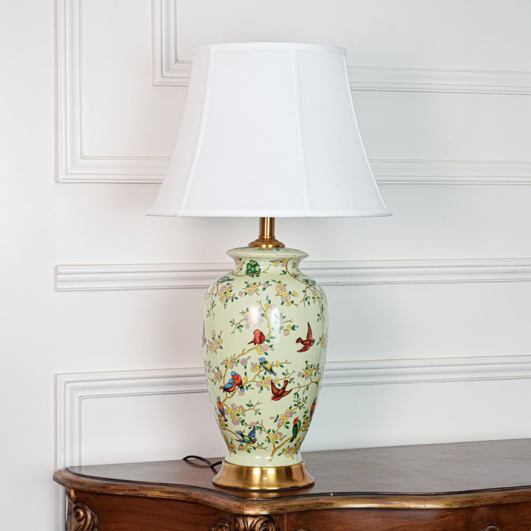 Decorative green ceramic table lamp with bird and floral artwork gold metal base and white shade on soft off white background.