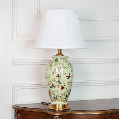 Decorative green ceramic table lamp with bird and floral artwork gold metal base and white shade on soft off white background.