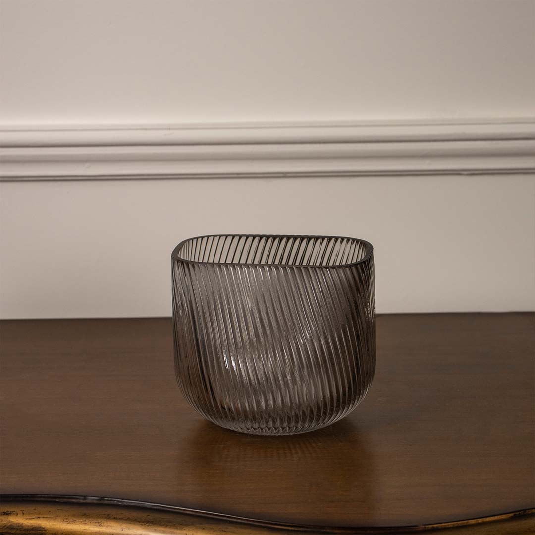 Smoky grey textured glass candle holder with ribbed finish placed on wooden table against neutral wall background for elegant ambient decor appeal.