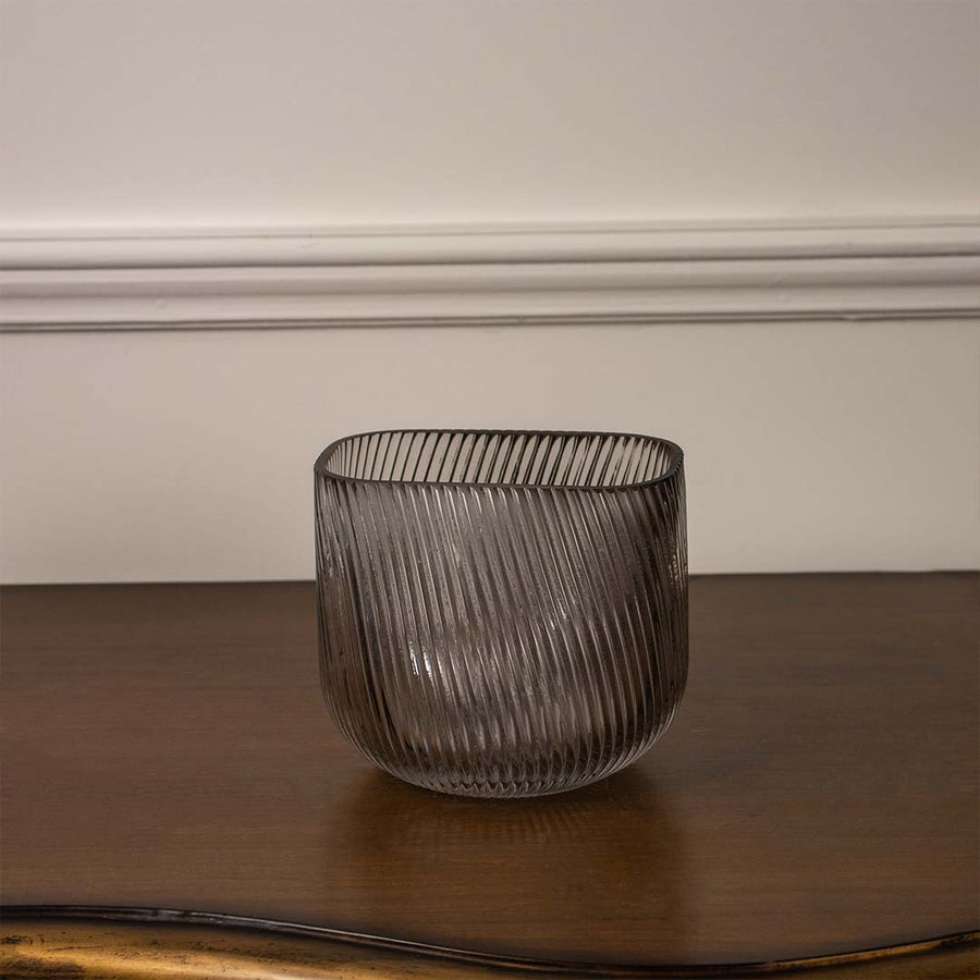 Smoky grey textured glass candle holder with ribbed finish placed on wooden table against neutral wall background for elegant ambient decor appeal.