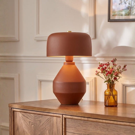 A terracotta-colored table lamp with a matte finish sits on a wooden console table next to a small amber glass vase with pink flowers, set against a cream-colored paneled wall.