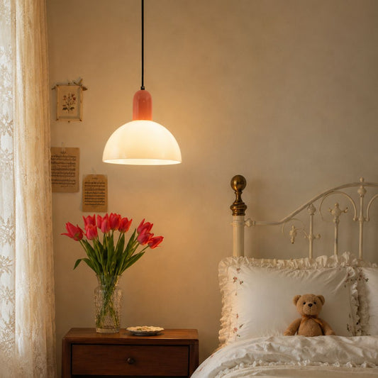 A warm, glowing pendant lamp with a white dome shade and a pinkish-orange cap hangs above a wooden nightstand in a cozy bedroom. A glass vase with red tulips sits on the table next to a bed with white ruffled bedding and a teddy bear.
