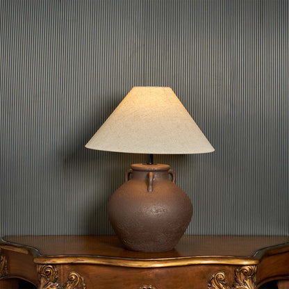 Brown ceramic table lamp with beige linen shade glowing on dark background styled as premium bedside table lamp.