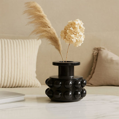 Black ceramic vase with raised spherical pattern placed on a marble coffee table with beige cushions in the background.