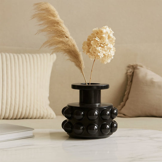 Black ceramic vase with raised spherical pattern placed on a marble coffee table with beige cushions in the background.