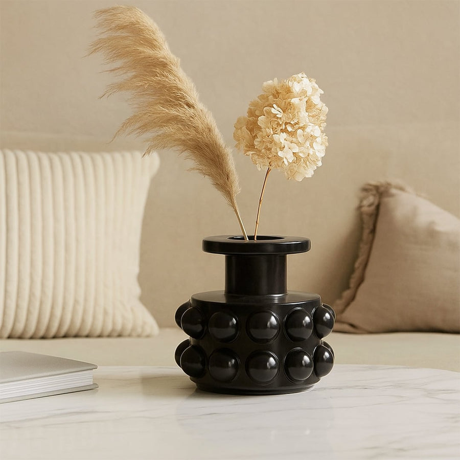 Black ceramic vase with raised spherical pattern placed on a marble coffee table with beige cushions in the background.