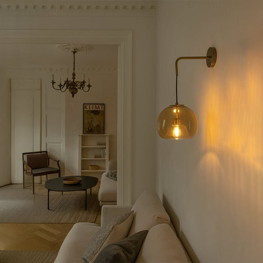 A warm living room scene with a glowing amber glass wall lamp casting soft golden light beside a beige sofa in a cozy modern interior style.
