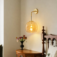Modern gold brass wall sconce featuring a glowing amber glass globe shade mounted in a cozy bedroom bedside corner.