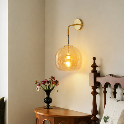 Modern gold brass wall sconce featuring a glowing amber glass globe shade mounted in a cozy bedroom bedside corner.