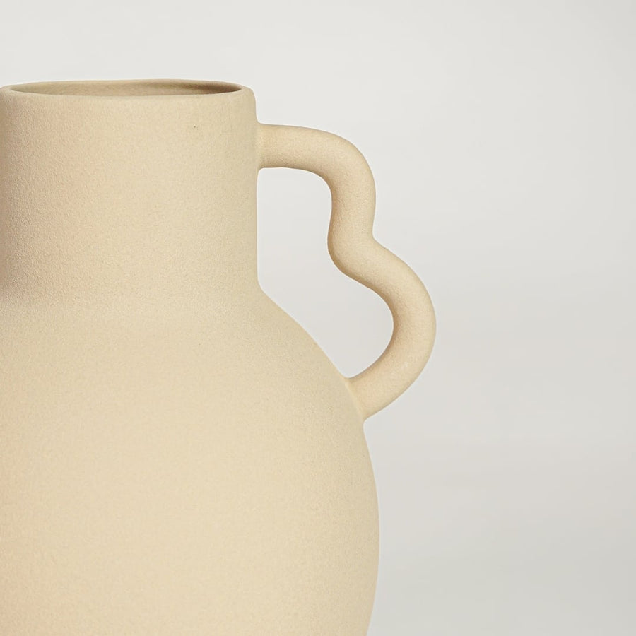 Closeup of matte beige ceramic vase with curved handles showcasing smooth finish ideal for shelf or side table decor.