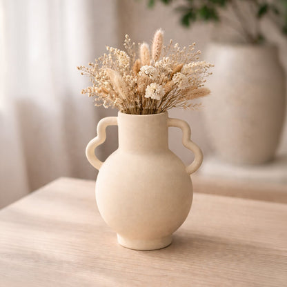 Beige ceramic double handle vase with dried stems placed on wooden table adding warm rustic charm to living room decor.