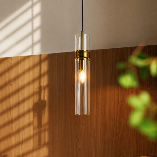 A modern cylindrical pendant light with a clear glass shade and brass metal accents, hanging against a warm wood-paneled wall with sunlight and window blinds casting shadows.