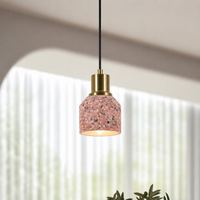 A modern pendant light with a terrazzo-patterned pink glass shade, a brass-finished metal collar, and a black hanging cord, shown in a softly lit interior setting.