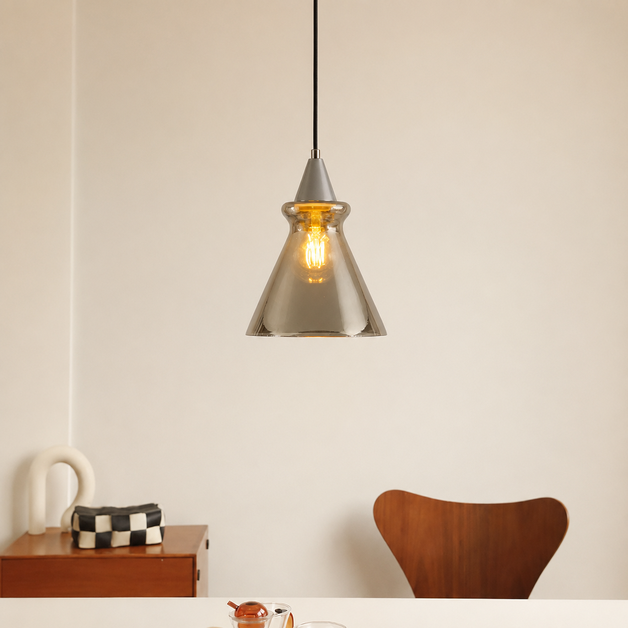 A modern pendant light with a brushed silver metal cone-shaped shade and a visible warm Edison bulb, hanging in a minimalist dining room setting above a table with a wooden chair and side cabinet.