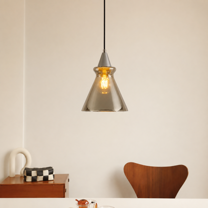 A modern pendant light with a brushed silver metal cone-shaped shade and a visible warm Edison bulb, hanging in a minimalist dining room setting above a table with a wooden chair and side cabinet.