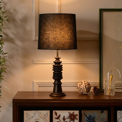 A table lamp with a dark brown textured fabric drum shade and a matching dark wood fluted base, placed on a wooden console table in a warmly lit room.