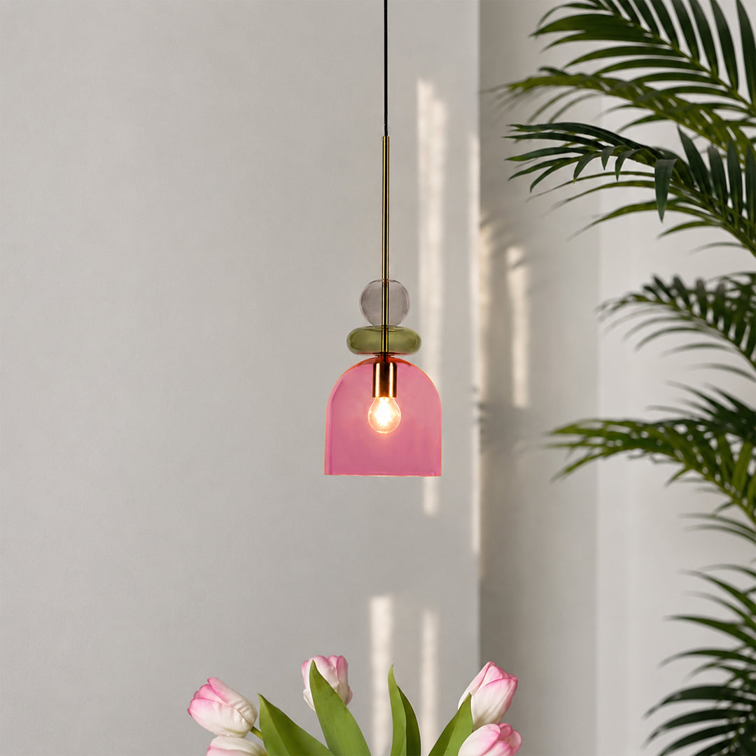A modern pendant light with a translucent pink glass dome shade, a brass rod, and stacked glass beads in green and clear, shown hanging in a minimalist room with tulips and a palm plant.