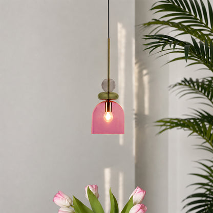 A modern pendant light with a translucent pink glass dome shade, a brass rod, and stacked glass beads in green and clear, shown hanging in a minimalist room with tulips and a palm plant.