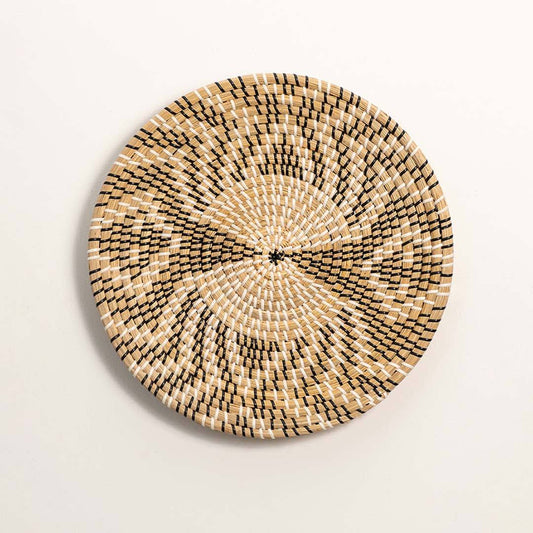 Round handcrafted black and natural seagrass wall basket with woven abstract geometric radial pattern on white background.
