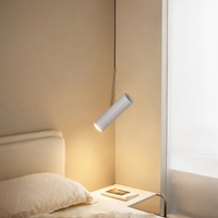 A modern cylindrical wall sconce lamp with a matte white finish, suspended by a thin black cord in a bedroom setting, casting a warm yellow glow next to a bed.