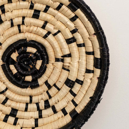 A closeup view of the natural and black rattan wall decor plate showing its textured spiral weaving and artisan craftsmanship for modern earthy home styling.