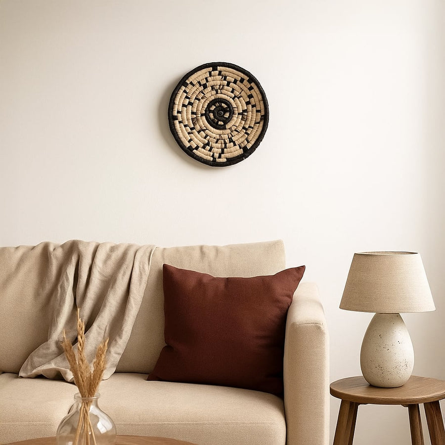 A handwoven natural and black rattan wall decor plate styled above a beige sofa with a brown cushion neutral throw and wooden side table creating warm cozy home vibes.