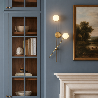 A brass wall sconce with two spherical frosted globe lights mounted on a dusty blue wall next to a framed landscape painting and above a white fireplace mantel.