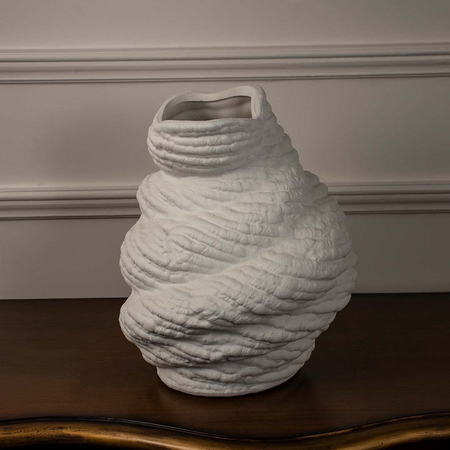 Twisted white ceramic vase with organic textured finish placed on wooden table against neutral wall background showcasing contemporary artistic home decor styling.