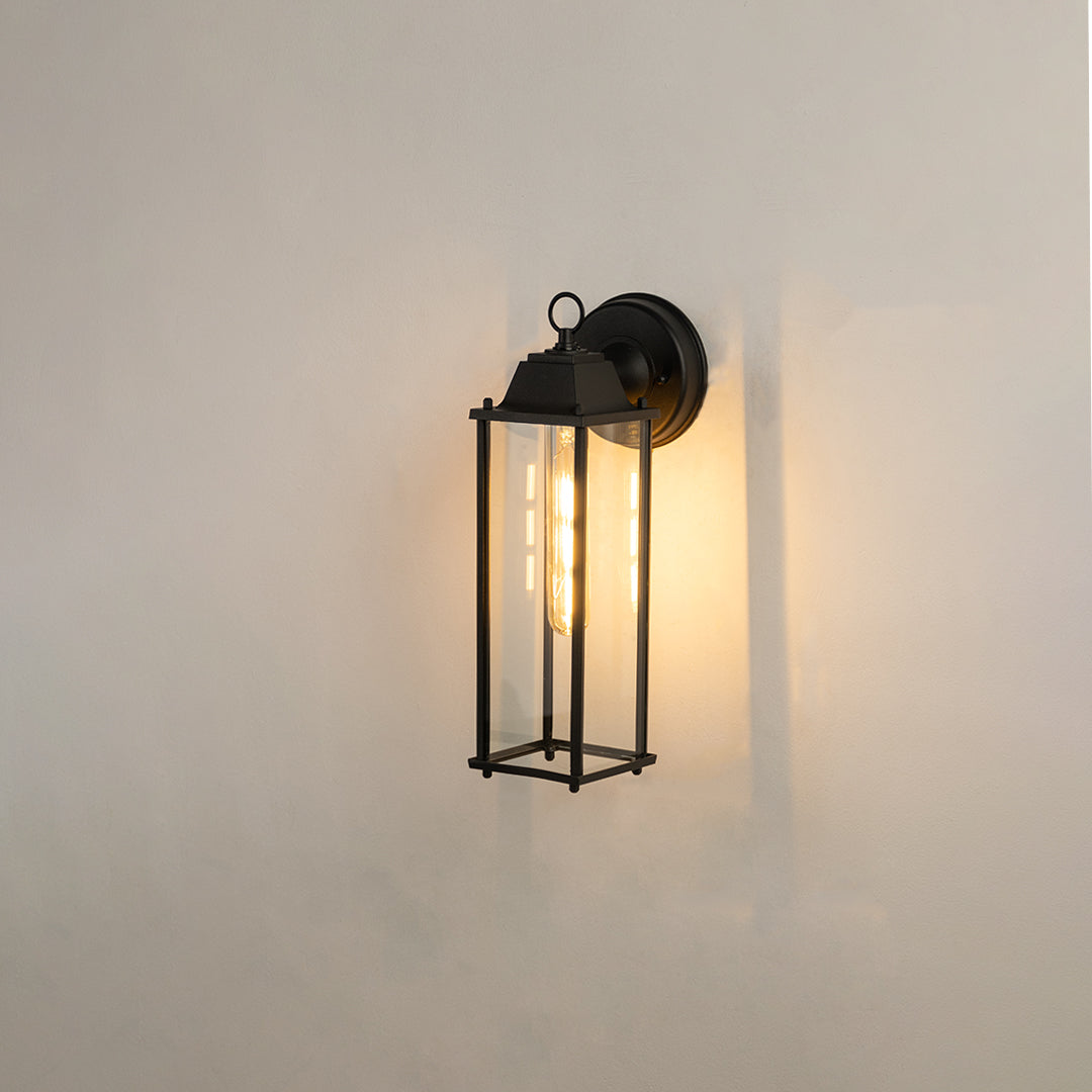 Minimalist black elongated outdoor wall light on white background with clear glass panels casting a warm downlight glow.