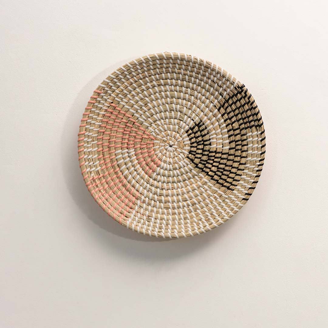 Round handcrafted natural seagrass wall basket with pink and black abstract color block patterns on white background.