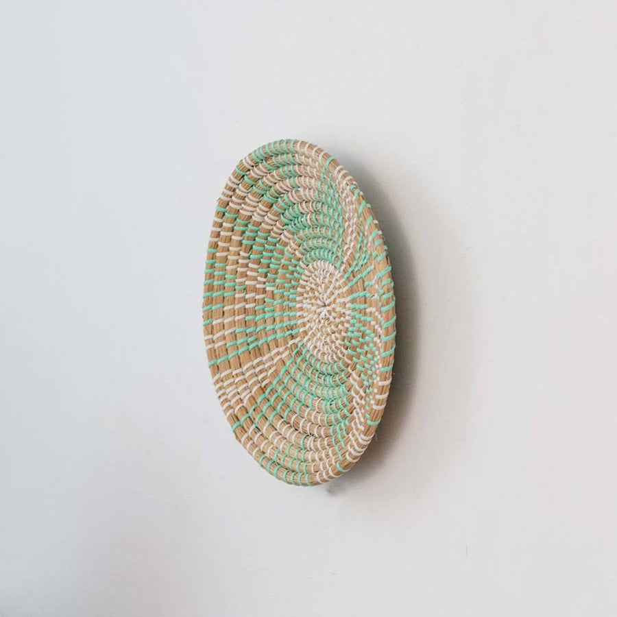 Handwoven seagrass round wall basket decor in natural beige and teal with raised spiral woven profile on white background.