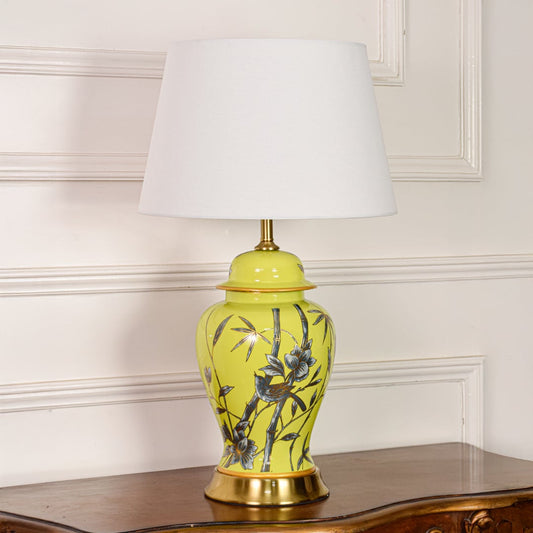 Decorative yellow ceramic table lamp with bamboo bird design gold metal base and fabric shade on off white backdrop.