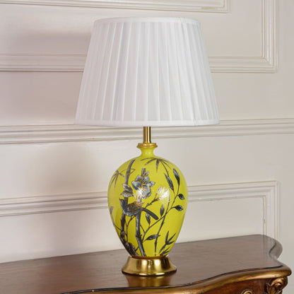 Decorative yellow ceramic table lamp with floral bird artwork gold metal base and pleated fabric shade on off white background.
