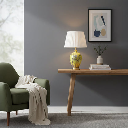 Yellow ceramic table lamp with hand painted floral bird artwork gold metal base and pleated fabric shade styled on side table.