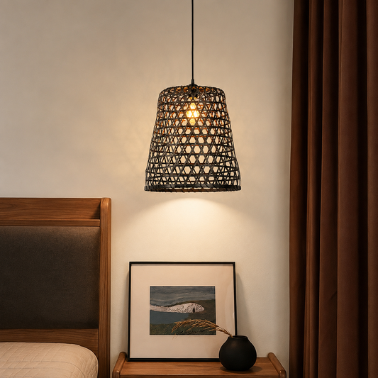 A woven black pendant light hangs over a bed in a bedroom setting, casting a warm glow. The scene includes a wooden headboard, a small framed landscape print, a black vase, and dark brown curtains.