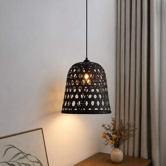 A black woven rattan pendant lampshade hanging in a room corner, casting a warm glow. The scene includes a beige wall, gray curtains, a wooden side table, and a small vase with dried flowers.