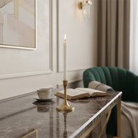 Slim gold candle holder with lit white taper candle placed on marble side table beside open book and cup creating calm elegant living room ambiance.