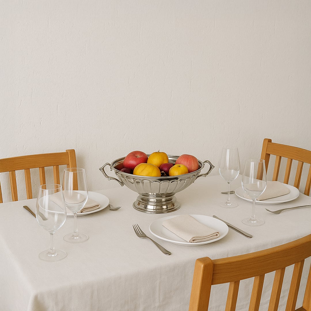 A silver fruit bowl filled with fresh apples and lemons placed at the center of a neatly set dining table with wooden chairs creating a warm home dining atmosphere.