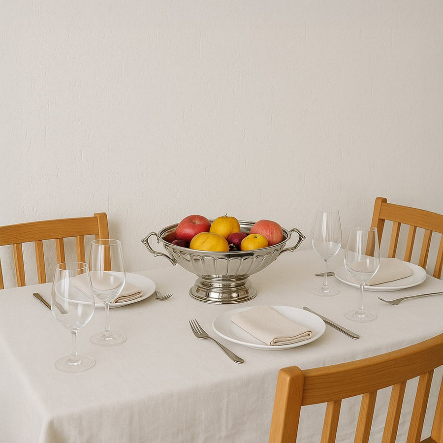 A silver fruit bowl filled with fresh apples and lemons placed at the center of a neatly set dining table with wooden chairs creating a warm home dining atmosphere.