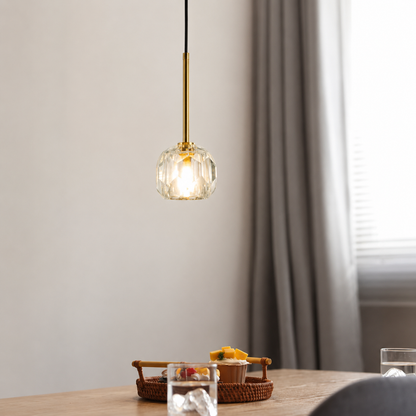 A modern pendant light with a clear faceted glass globe shade and a brushed gold metal rod, hanging over a wooden table in a softly lit room with gray curtains.