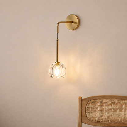 A modern wall sconce with a brushed gold metal arm and circular wall mount, featuring a clear faceted glass shade that glows with warm light, shown mounted on a beige wall above a wooden chair.