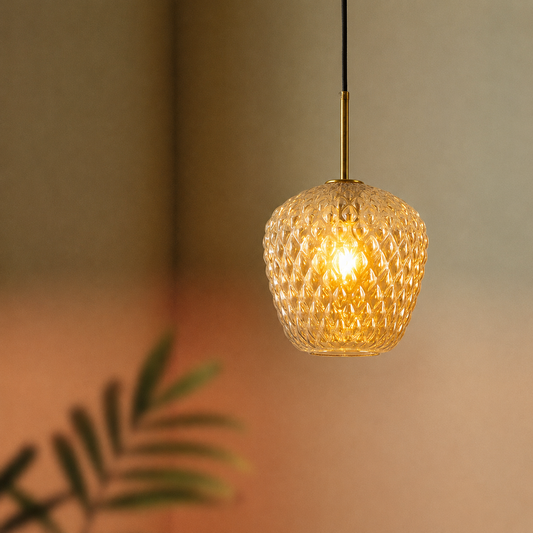 A warm glowing pendant light with a textured, honeycomb-like glass shade suspended by a slim brass rod against a softly blurred beige wall with a hint of green foliage in the foreground.