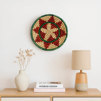 Handwoven round wall basket Decor in green red and natural tones styled above a wooden console with ceramic vases table lamp and books creating a warm minimalist home setting.