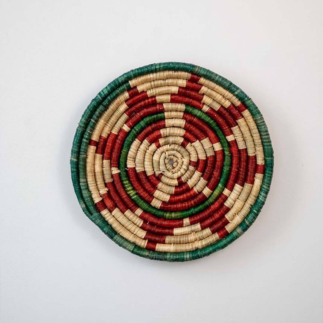 Handwoven woven wall basket decor in natural red green and beige with geometric spiral pattern displayed on white background.