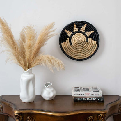 Handcrafted round woven wall basket styled above wooden console with ceramic vases and dried grass creating warm boho inspired living room wall decor.