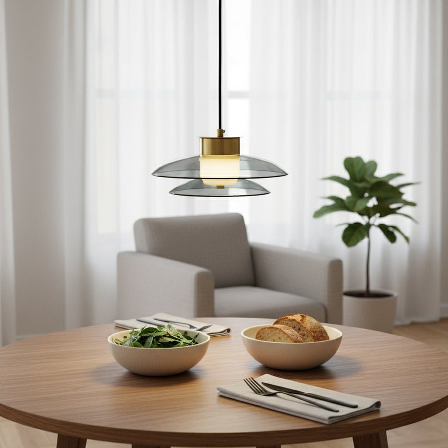 Modern grey glass pendant light with gold metal fitting glowing warmly above a round dining table creating a soft cozy atmosphere in a minimal living space.