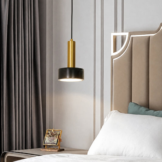 A modern pendant light with a black cylindrical shade and a gold cylindrical accent, hanging in a styled bedroom next to gray curtains and a beige upholstered headboard.